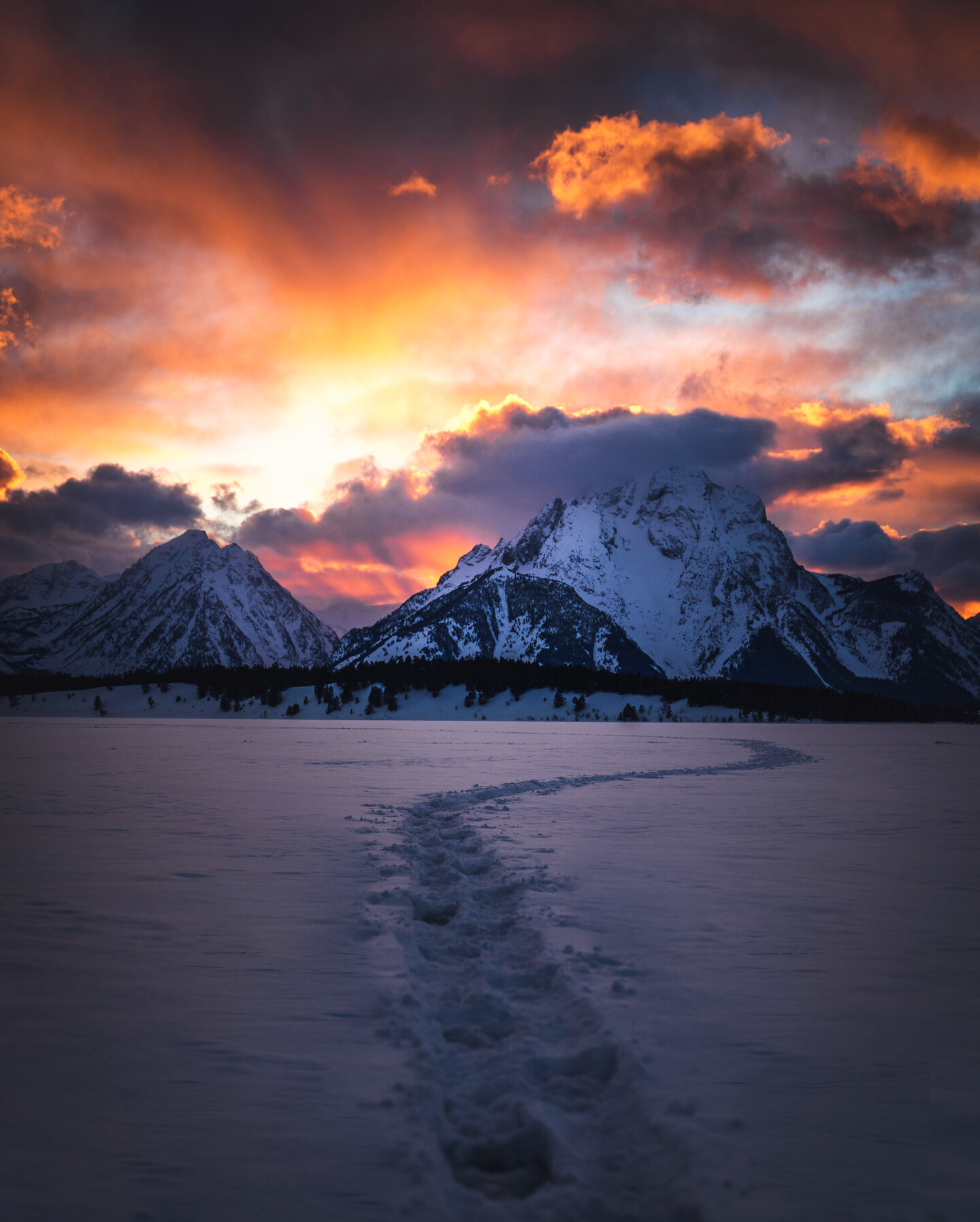 Grand Tetons at sunset — snow-covered foreground with a single set of footprints leading through the snow toward the peaks, fiery orange and red clouds breaking over the range.