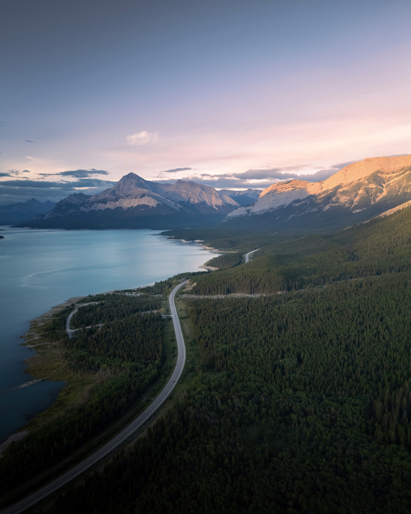 Aerial view of a winding mountain road along a glass-still lake, Canadian Rockies in the distance carrying pink-peach alpenglow on the peaks under a pale dawn sky.