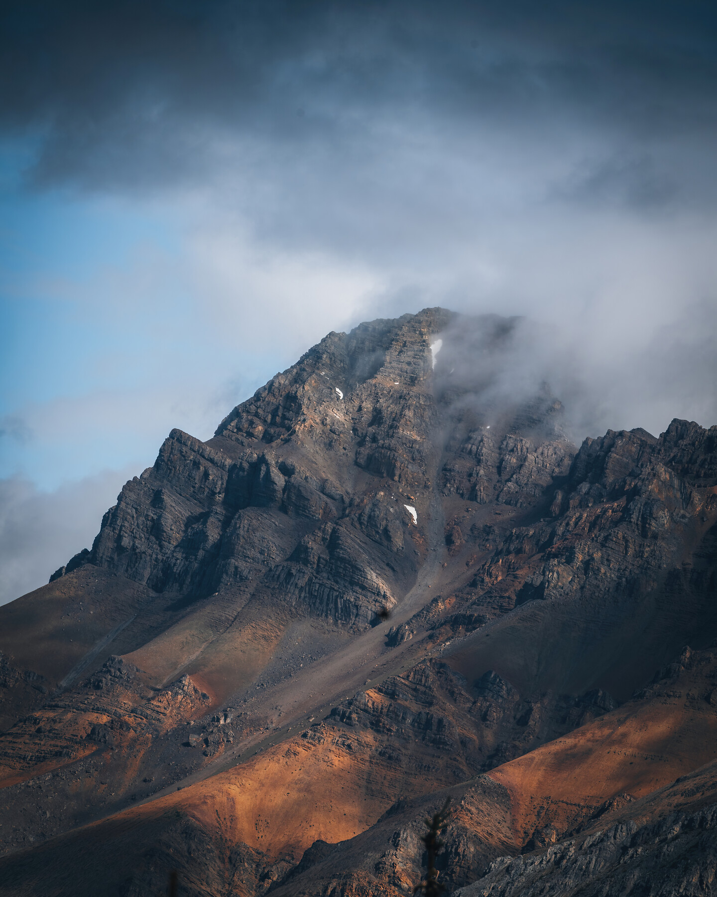 A jagged alpine peak wrapped in cloud — warm-lit lower slopes giving way to a fog-shrouded summit, light breaking through atmospheric weather.