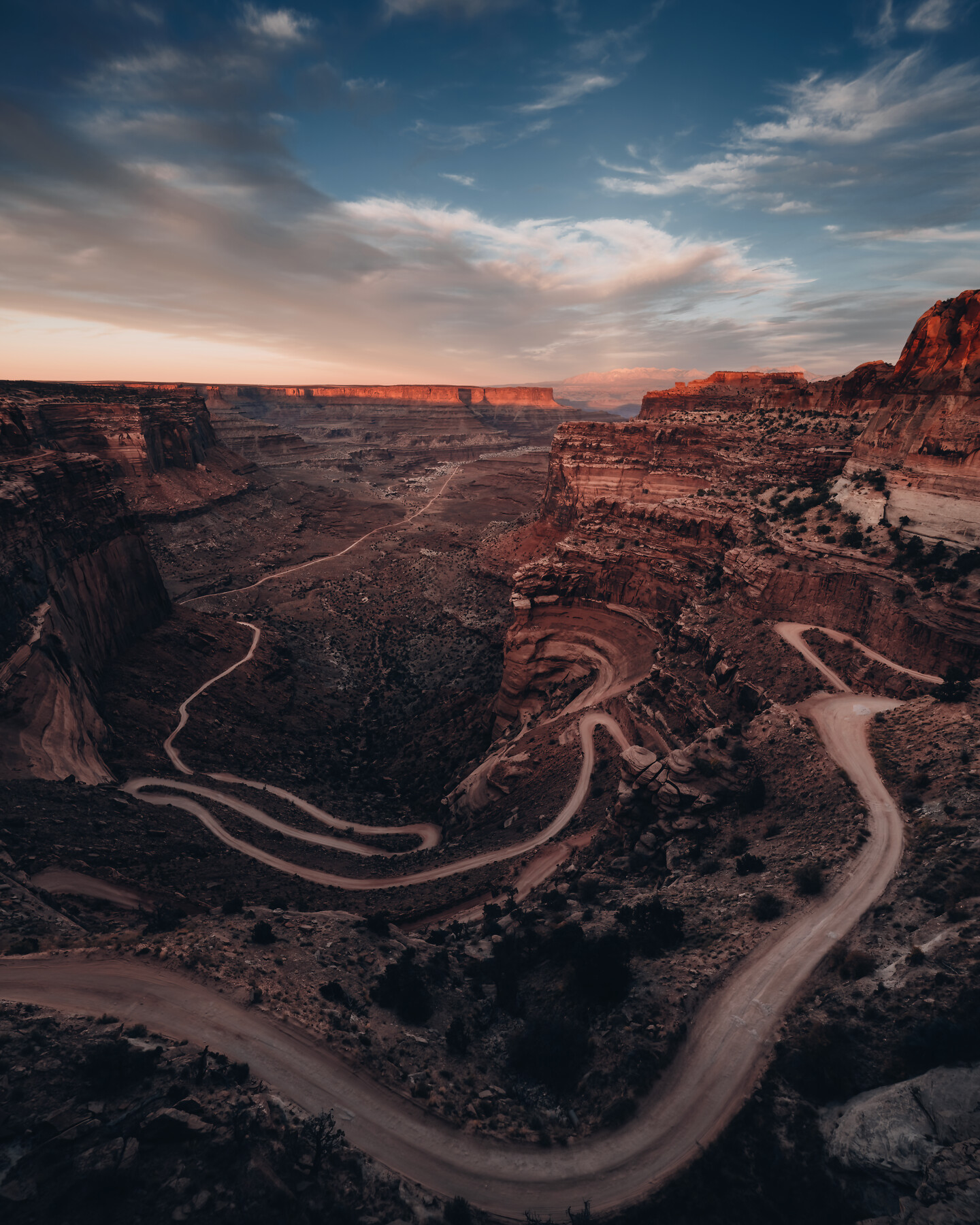 Aerial view of a switchback dirt road carved into red Canyonlands cliffs at dusk — pink alpenglow on the far canyon walls under a streaked blue sky, deep canyon shadows below.