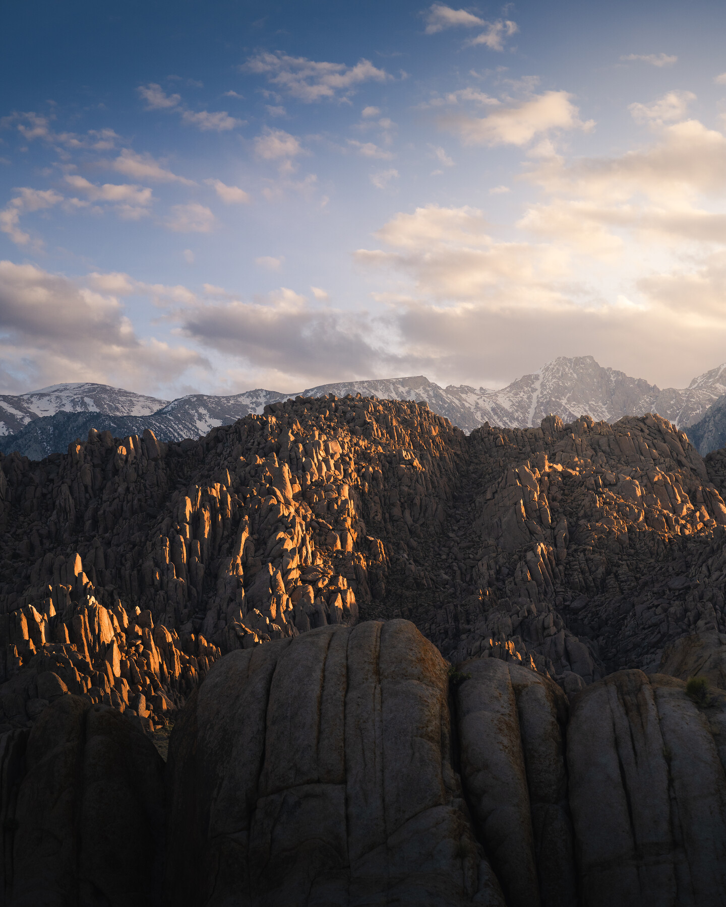 Eastern Sierra rocky pinnacles at sunset — warm rim-light catching textured rock formations, snow-capped peaks behind, pale blue sky above.