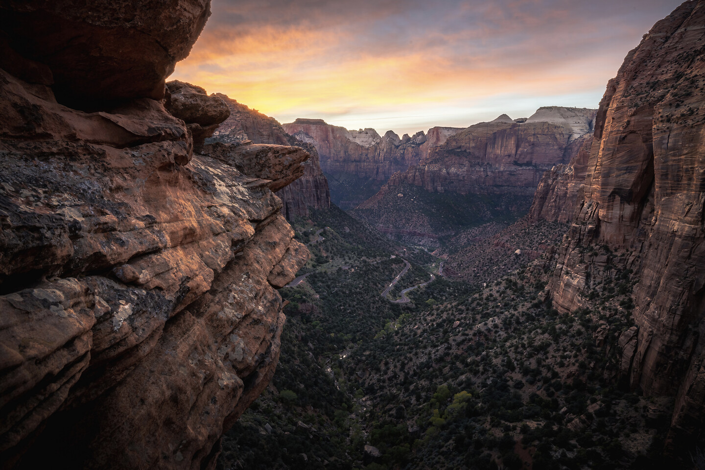Zion canyon viewed from a high overlook at sunset — red sandstone cliffs framing a sweeping valley, warm orange and pink light over the canyon, soft layered clouds.