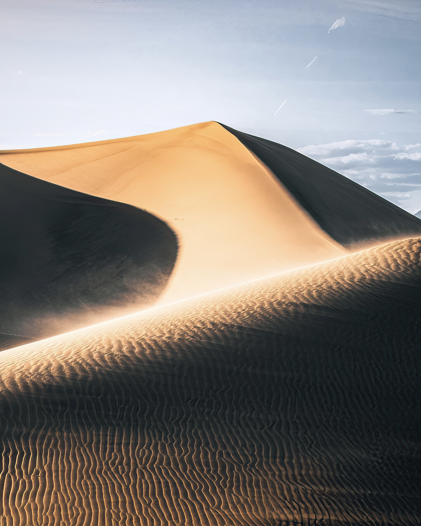 A Death Valley sand dune sculpted by side-light — a strong shadow line bisects the dune crest, warm-lit ripples leading down into the foreground, pale blue sky.