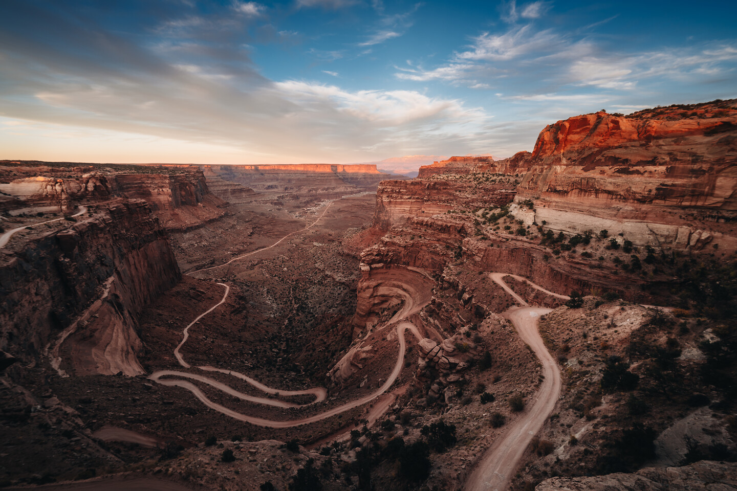 Aerial sweep of the Shafer Canyon switchback road in Canyonlands — the dirt road S-curves through red rock cliffs at sunset, dramatic streaked sky above.