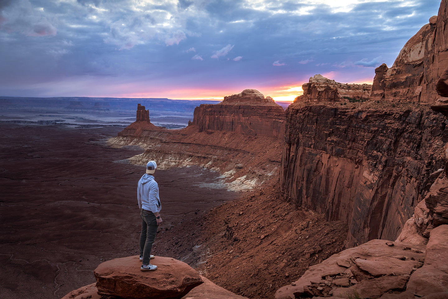 A lone figure stands on a rock ledge above a deep canyon at twilight — distant mesas catching the last band of sunset, cool blue dusk settling over the canyon walls.